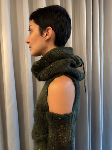 Person wearing a dark green knitted vest against a neutral curtain background